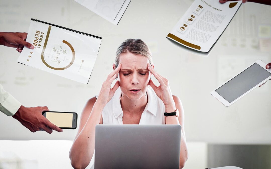 Mulher de negócios estressada, sentada em frente a um laptop, sobrecarregada com papéis e dispositivos eletrônicos.